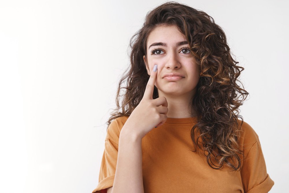 young woman with long, curly brown hair wearing an orange shirt points to a blemish or spot on her cheek with a concerned or unhappy expression, suggesting a skin issue.