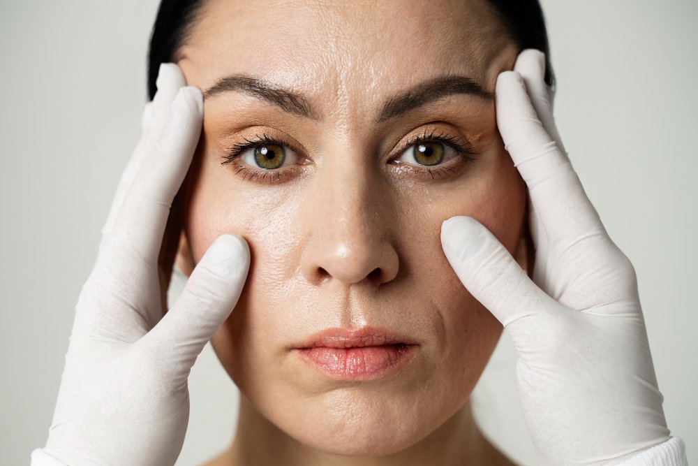 Close-up of a woman's face with a neutral expression, being gently touched on the temples and cheeks by hands wearing white medical gloves, suggesting a dermatological or cosmetic procedure.